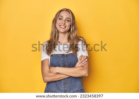 Young blonde Caucasian woman in denim overalls posing on a yellow background, who feels confident, crossing arms with determination.