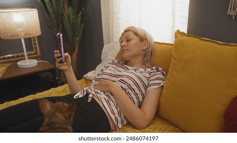 A young blonde caucasian woman relaxing on a bed in a cozy bedroom with her chihuahua dog, checking her phone indoors at home.