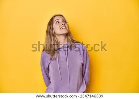 Young blonde Caucasian woman in a violet sweatshirt on a yellow background, relaxed and happy laughing, neck stretched showing teeth.