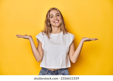 Young blonde Caucasian woman in a white t-shirt on a yellow studio background, makes scale with arms, feels happy and confident. - Powered by Shutterstock