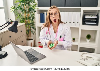 Young Blonde Doctor Woman Holding Pills In Shock Face, Looking Skeptical And Sarcastic, Surprised With Open Mouth 