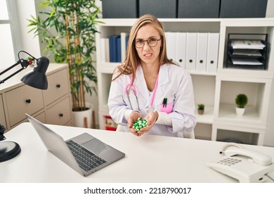 Young Blonde Doctor Woman Holding Pills Clueless And Confused Expression. Doubt Concept. 