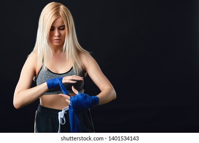 Young blonde female boxer wrapping his hands with blue elastic bandage preparing to workout on black background - Powered by Shutterstock