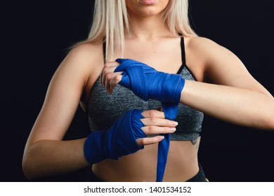 Young blonde female boxer wrapping his hands with blue elastic bandage preparing to workout on black background - Powered by Shutterstock