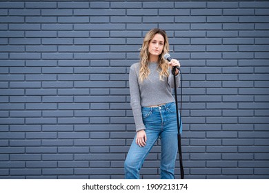 Young Blonde Female Comedian Against Blue Grey Brick Wall With Microphone And Expressions 