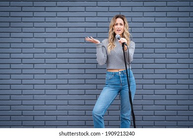 Young Blonde Female Comedian Against Blue Grey Brick Wall With Microphone And Expressions 