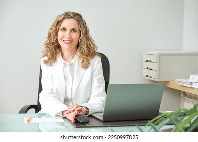 young blonde female doctor in white coat smiling at the table in her office looking at the camera with the computer next to her - Powered by Shutterstock