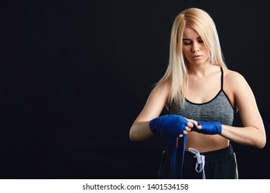 Young blonde female Muay Thai fighter wrapping his hands with blue elastic bandage preparing to workout on black background