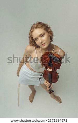 a young blonde female musician stands with a violin in one hand and a bow in the other top view