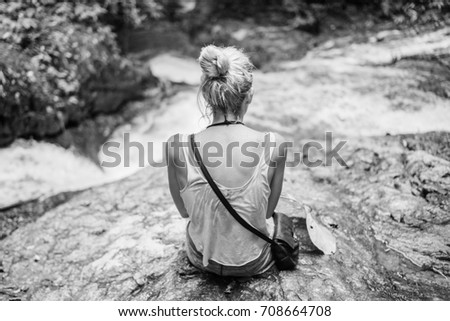 Young blonde female sitting on the cliff at the waterfall. Hiking travel freedom concept. 