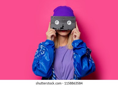 Young blonde girl in 90s sports jacket and VHS cassette on pink background.