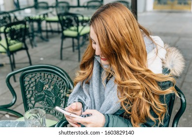 Young Blonde Girl With Coat Smiles While Checking Her Smartphone Sitting In A Bar Having A Drink