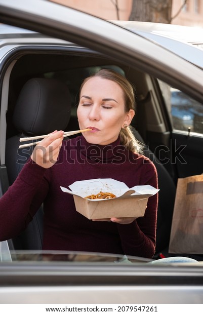 Young\
blonde girl eating delicious Chinese noodles from the box with\
chopsticks, sitting in the car. Wok noodle\
concept