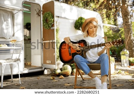 A young blonde girl with a guitar in front of a travel trailer. Rest outside the city. Happy people enjoy weekends in the summer.