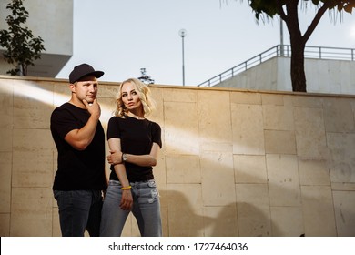 
A Young Blonde Girl And Her Boyfriend Are Standing Next To A Light Wall In A Ray Of Light. Dressed In Black T-shirts And Jeans.