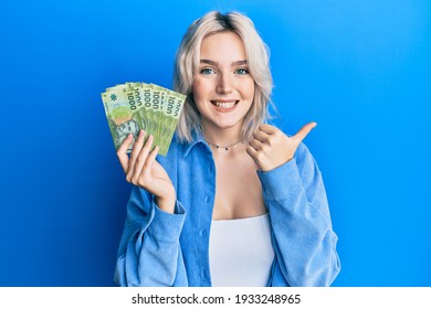 Young blonde girl holding 1000 chilean pesos pointing thumb up to the side smiling happy with open mouth 