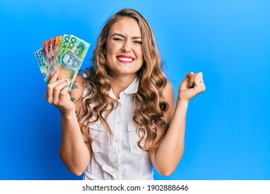 Young blonde girl holding australian dollars screaming proud, celebrating victory and success very excited with raised arm 