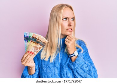 Young Blonde Girl Holding Canadian Dollars Thinking Worried About A Question, Concerned And Nervous With Hand On Chin 