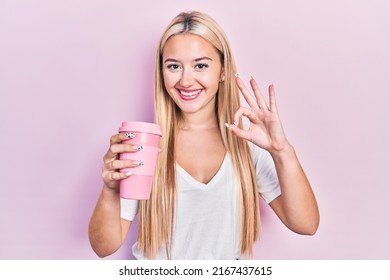 Young Blonde Girl Holding Coffee Doing Ok Sign With Fingers, Smiling Friendly Gesturing Excellent Symbol 