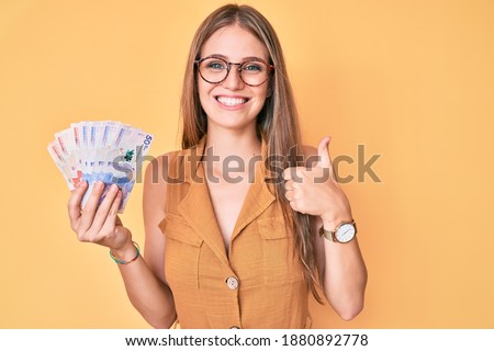 Young blonde girl holding colombian pesos smiling happy and positive, thumb up doing excellent and approval sign 