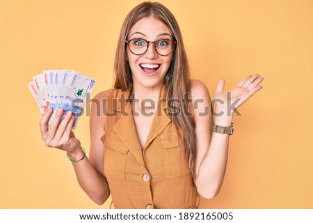 Young blonde girl holding colombian pesos celebrating victory with happy smile and winner expression with raised hands 