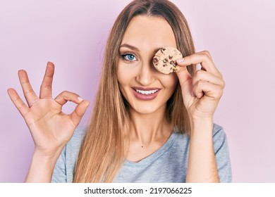 Young Blonde Girl Holding Cookie Over Eye Doing Ok Sign With Fingers, Smiling Friendly Gesturing Excellent Symbol 