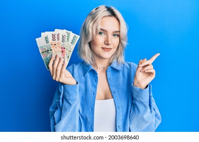 Young blonde girl holding czech koruna banknotes smiling happy pointing with hand and finger to the side 