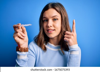 Young Blonde Girl Holding Dental Orthodontic Clear Aligner Over Blue Isolated Background Surprised With An Idea Or Question Pointing Finger With Happy Face, Number One