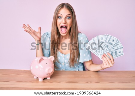Young blonde girl holding dollars and piggy bank sitting on the table celebrating crazy and amazed for success with open eyes screaming excited. 