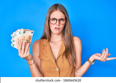 Young Blonde Girl Holding Euro Banknotes Clueless And Confused Expression. Doubt Concept. 