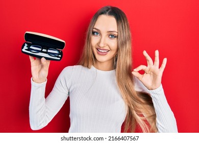 Young Blonde Girl Holding Glasses Doing Ok Sign With Fingers, Smiling Friendly Gesturing Excellent Symbol 