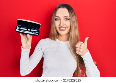 Young Blonde Girl Holding Glasses Smiling Happy And Positive, Thumb Up Doing Excellent And Approval Sign 