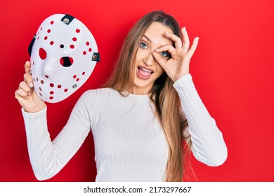 Young Blonde Girl Holding Hockey Mask Smiling Happy Doing Ok Sign With Hand On Eye Looking Through Fingers 