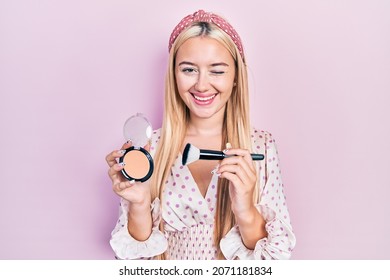 Young blonde girl holding makeup bronzer and brush winking looking at the camera with sexy expression, cheerful and happy face. 