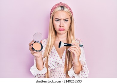 Young blonde girl holding makeup bronzer and brush skeptic and nervous, frowning upset because of problem. negative person. 