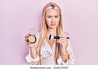 Young blonde girl holding makeup bronzer and brush clueless and confused expression. doubt concept. 