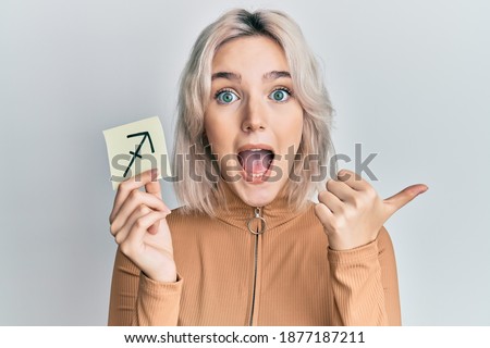 Young blonde girl holding paper with sagittarius zodiac sign pointing thumb up to the side smiling happy with open mouth 
