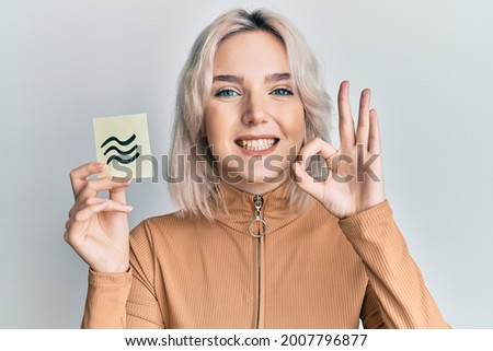 Young blonde girl holding paper with aquarius zodiac sign doing ok sign with fingers, smiling friendly gesturing excellent symbol 