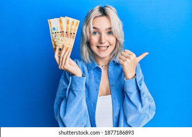 Young blonde girl holding philippine peso banknotes pointing thumb up to the side smiling happy with open mouth 