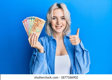 Young blonde girl holding philippine peso banknotes smiling happy and positive, thumb up doing excellent and approval sign 