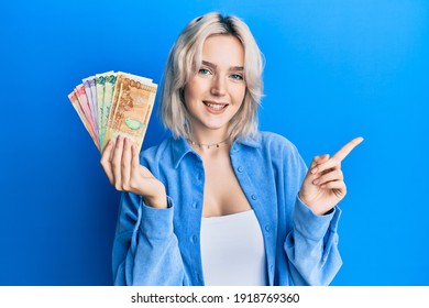 Young blonde girl holding philippine peso banknotes smiling happy pointing with hand and finger to the side 