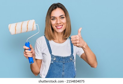 Young Blonde Girl Holding Roller Painter Smiling Happy And Positive, Thumb Up Doing Excellent And Approval Sign 