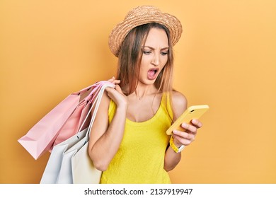 Young Blonde Girl Holding Shopping Bags Using Smartphone Angry And Mad Screaming Frustrated And Furious, Shouting With Anger. Rage And Aggressive Concept. 