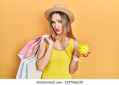 Young Blonde Girl Holding Shopping Bags And Piggy Bank Clueless And Confused Expression. Doubt Concept. 