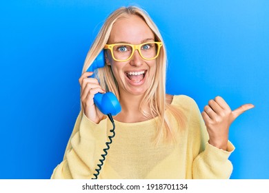 Young blonde girl holding vintage telephone pointing thumb up to the side smiling happy with open mouth 