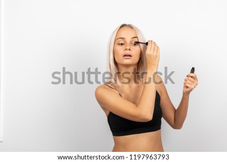Young blonde girl paints her eyelashes with black mascara on a white background