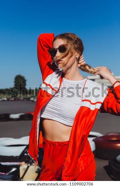 Young blonde girl in red sports suit and\
sunglasses posing in summer on a kart\
track