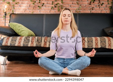 young blonde girl sitting on the wooden floor in the living room with her legs crossed and her eyes closed meditating