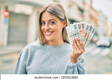 Young blonde girl smiling happy holding czech koruna banknotes at the city.