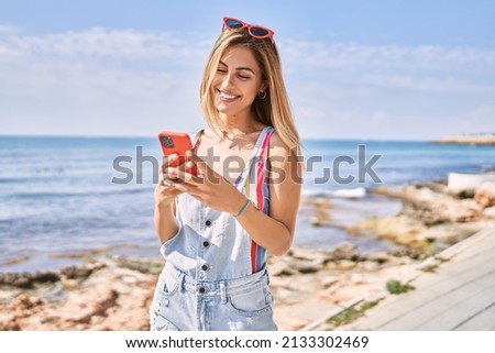 Young blonde girl smiling happy using smartphone at the beach.